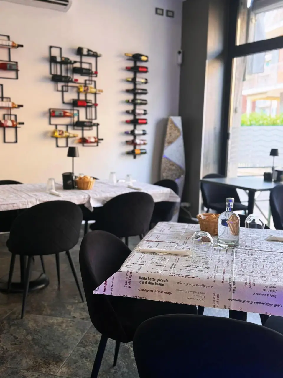 La sala con i tavoli apparecchiati, le tovagliette di carta a giornale e le rastrelliere dei vini